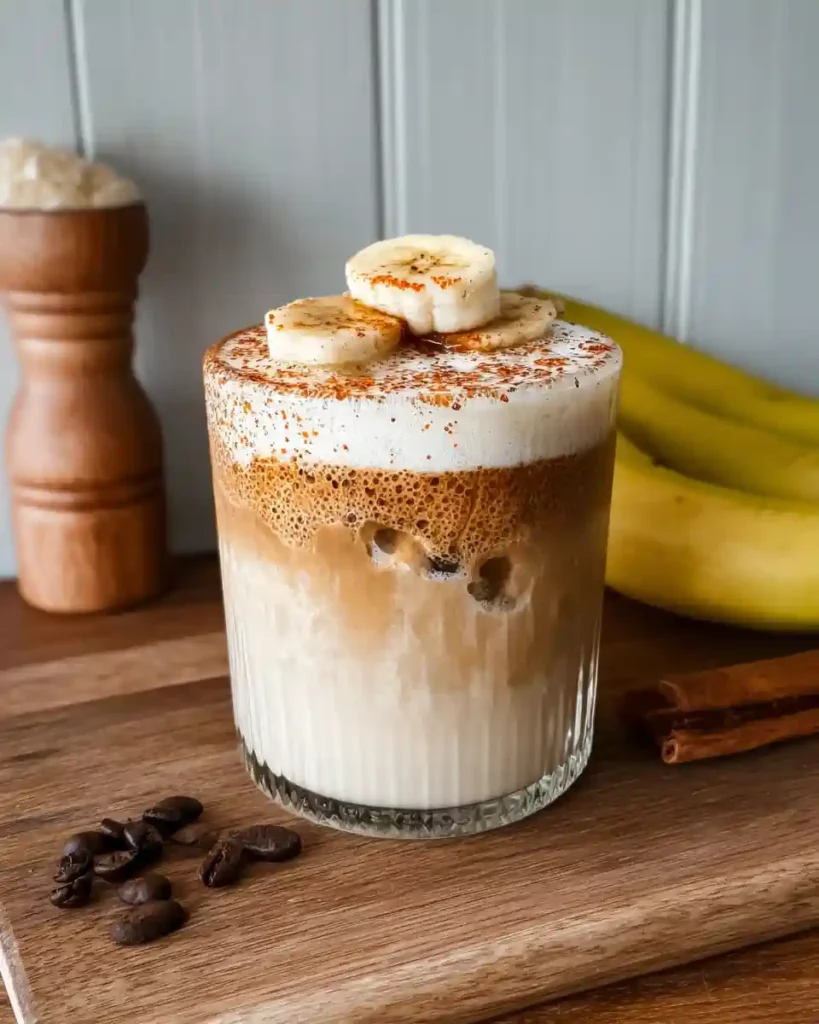 How to Make a Cozy Banana Bread Latte at Home 5 Iced banana latte with frothy milk foam and banana slices on top, served on a wooden board with coffee beans and cinnamon sticks